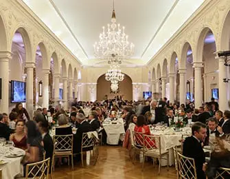 Grand ballroom with arched colonnades, ornate chandeliers, and tables set for a formal event with many guests.