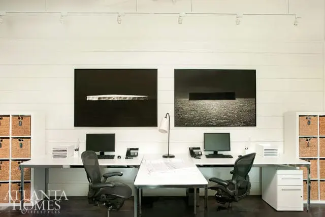 Modern office with white shiplap walls, two white desks with computers and black ergonomic chairs, and two framed abstract art pieces depicting icebergs.
