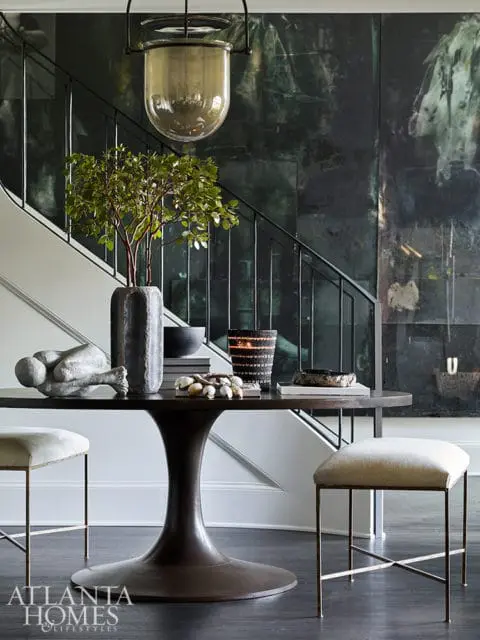 Dark wood tulip pedestal table with decorative objects, two upholstered stools, and a large abstract artwork on a dark green wall with a staircase in the background.