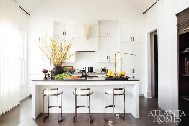 White kitchen with white cabinetry, dark countertop island, three brass and white bar stools, and a white bull head mounted above the range hood.