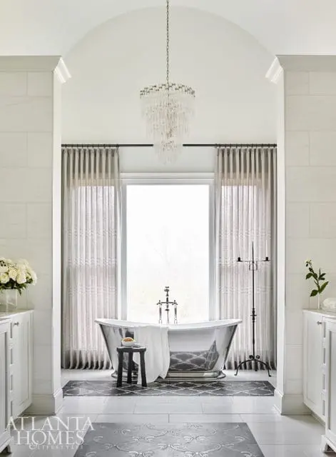 White bathroom with a freestanding silver bathtub, crystal chandelier, floor-to-ceiling marble tile, and patterned floor tiles.