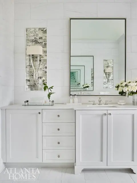 White bathroom vanity with marble tile walls, a large framed mirror, and decorative sconces.