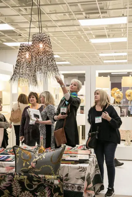 Women examining a display of decorative pillows and fabric samples at a trade show, with a woman reaching up to touch a unique pendant light fixture.