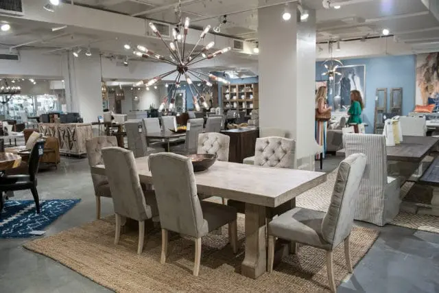 Dining room set with a light wood table and upholstered chairs in neutral tones, under a starburst chandelier in a showroom.
