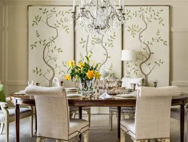 Dining room with a dark wood table set for a meal, upholstered chairs, a crystal chandelier, and a triptych wall panel featuring a painted tree with birds.