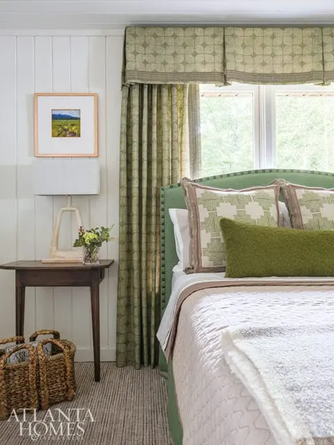 Bedroom with green upholstered headboard, patterned green curtains, white paneled walls, and a small wooden side table with a lamp and basket.