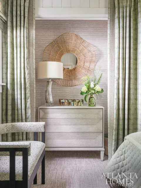 Bedroom with light wood dresser, woven sunburst mirror, upholstered chair, and patterned curtains.