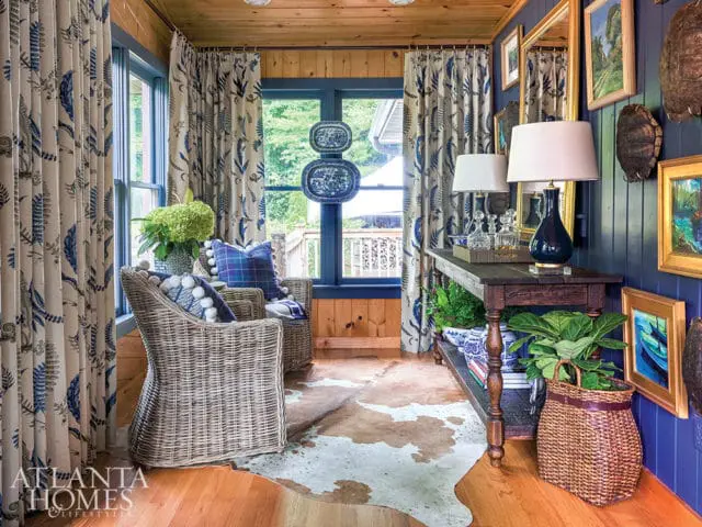 Sunroom with wicker chairs, blue and white botanical print curtains, dark wood console table with lamps, and blue paneled walls adorned with artwork and tortoise shells.