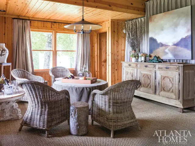 Living room with wood-paneled walls and ceiling, featuring wicker chairs around a round table, a rustic wooden sideboard, and a large abstract painting.