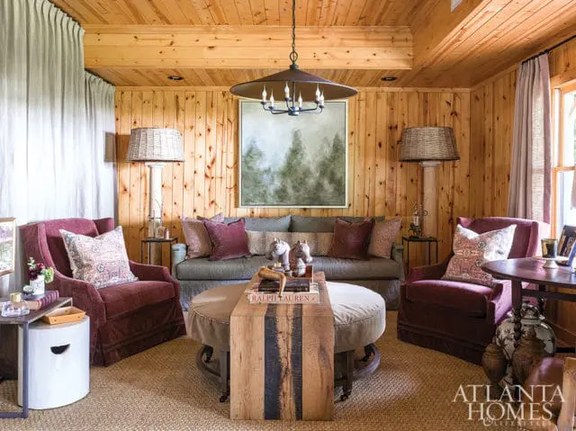 Living room with wood-paneled walls and ceiling, featuring plush burgundy armchairs, a gray sofa with patterned pillows, a large round ottoman coffee table, and a landscape painting above the sofa.