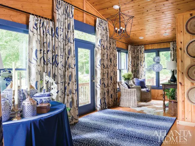 Sunroom with wood-paneled walls and ceiling, featuring blue and white botanical print curtains, wicker armchairs, a blue patterned rug, and a table with blue and white ginger jars.
