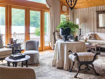 Living room with wood-paneled ceiling, patterned curtains, and upholstered furniture including a sofa, armchairs, and a bench. A round table with a large plant sits center.
