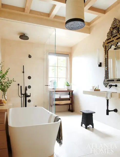 Cream tiled bathroom with a freestanding white tub, glass-enclosed shower with black fixtures, stone vanity, and ornate black mirror above the sink.