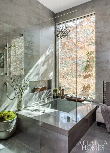 Modern bathroom with a grey tiled shower enclosure, a built-in soaking tub, and a large window overlooking trees with fall foliage.