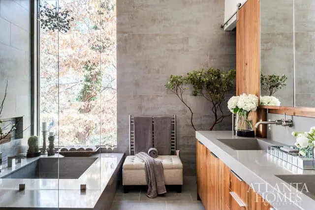 Modern bathroom with a large concrete soaking tub, grey tiled walls, wooden vanity with double sinks, and a large window overlooking trees.