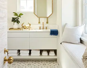 Modern bathroom vanity with white drawers and open shelving displaying white towels and soaps. Features a white countertop, gold faucet, hexagonal mirror, and a gold crab sculpture.