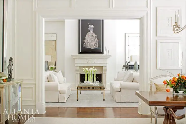 Living room with two white slipcovered sofas flanking a marble fireplace, a glass coffee table with calla lilies, and a framed artwork above the mantel.