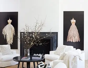 Living room with two white armchairs flanking a black fireplace, each with a large framed artwork of a dress on a mannequin above. A round black coffee table sits in front of the fireplace.