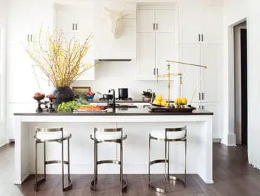 White kitchen with white Shaker cabinets, black granite island, three white and brass bar stools, and a decorative bull head mounted above the range hood.