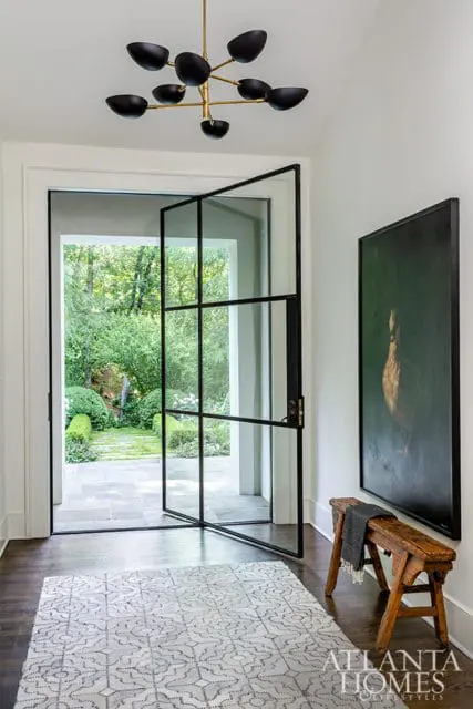 Entryway with a black metal-framed glass door opening to a garden, a modern brass chandelier, a framed painting of a bird, a wooden bench, and a patterned rug.