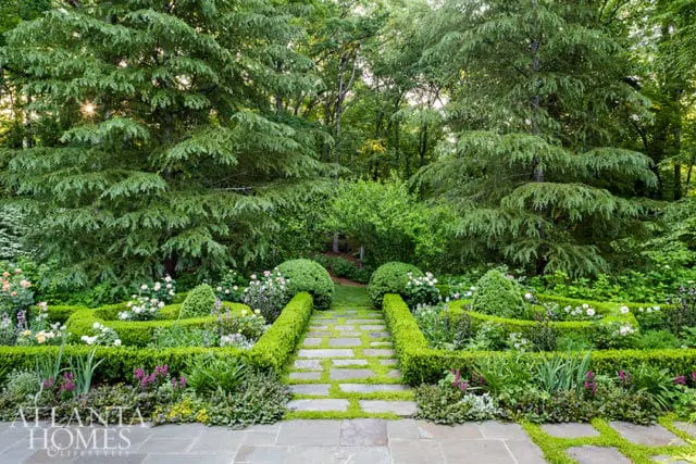 Stone pathway with grass between the stones leading through a formal garden with manicured hedges, roses, and lush trees.