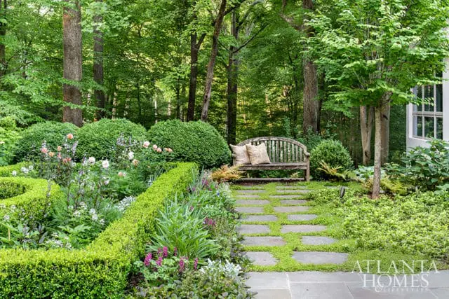 Stone pathway through a lush garden with a wooden bench, manicured hedges, and blooming flowers, leading towards a wooded area.