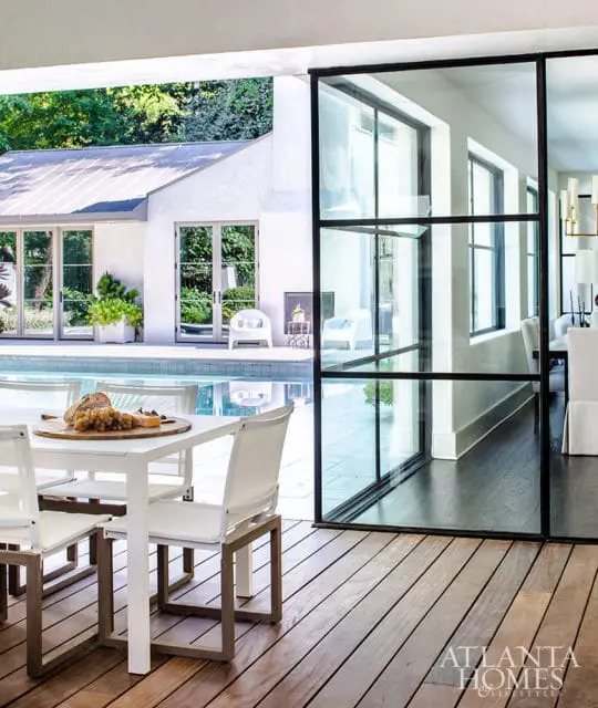 White dining set with chairs on a wooden deck overlooking a pool and modern interior with large glass doors