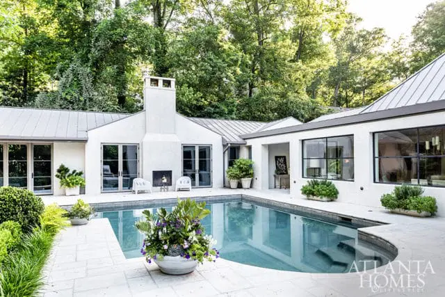 Modern white stucco home with a metal roof surrounding a freeform swimming pool, featuring a tall chimney and large windows. Lush greenery and potted plants accent the patio.