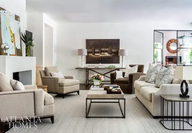 Living room with neutral-toned furniture, including a fireplace, sofas, armchairs, and a coffee table. Abstract art hangs above the fireplace and a landscape painting is on the wall.