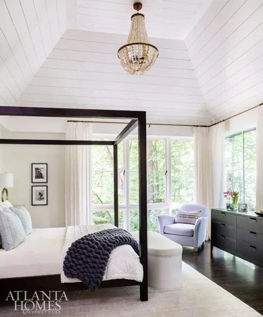 Bedroom with a dark wood four-poster bed, white shiplap vaulted ceiling, beaded chandelier, light blue armchair, and dark dresser.