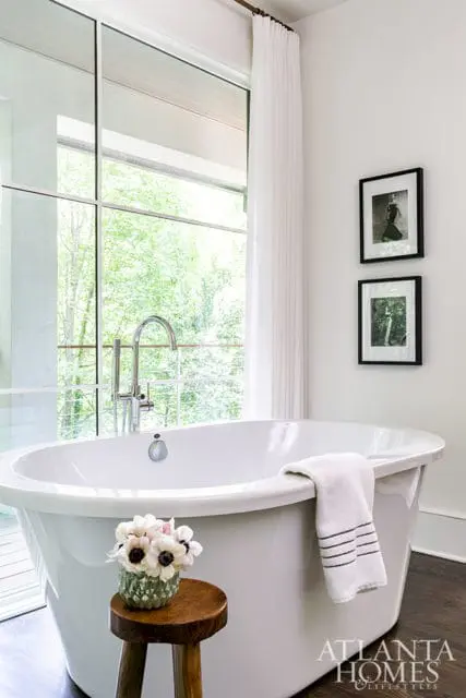 White freestanding bathtub with chrome faucet, next to a large window overlooking trees. Two framed black and white photos hang on the wall.