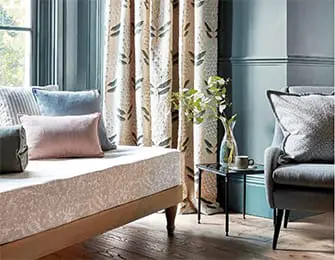 Daybed with patterned upholstery and pastel throw pillows next to a side table with a vase of greenery and a coffee cup.
