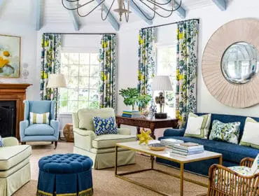 Living room with blue sofa, light green armchairs, and blue tufted ottoman. Features floral curtains, a wooden fireplace, and a large round mirror above the sofa.