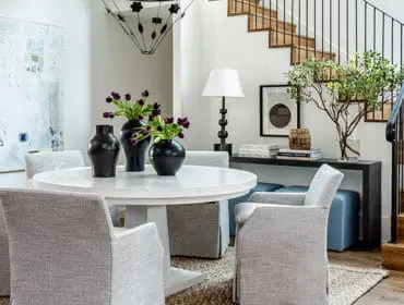 Dining area with a white pedestal table, upholstered chairs, black vases with purple tulips, and a modern chandelier. A wooden staircase with black railings is visible in the background.