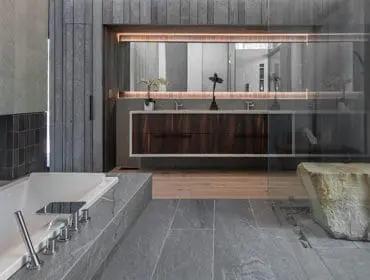 Modern bathroom with a white soaking tub, grey stone tile flooring, dark wood vanity with dual sinks, and a large rock feature in the glass-enclosed shower.