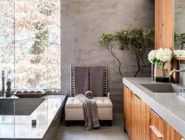 Modern bathroom with concrete bathtub, wood cabinetry, gray tile walls, and a large window overlooking trees.