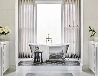 White bathroom with freestanding clawfoot tub, patterned rug, and dual vanities with white cabinetry. Large window with sheer curtains behind the tub.