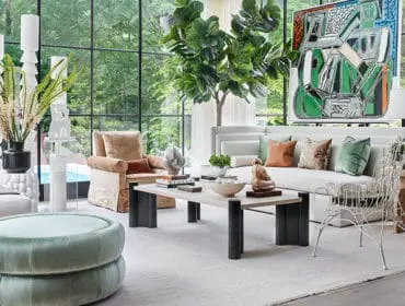 Living room with large windows overlooking greenery, featuring a white sectional sofa with colorful pillows, a tan armchair, a light wood coffee table with sculptures, and a mint green ottoman.