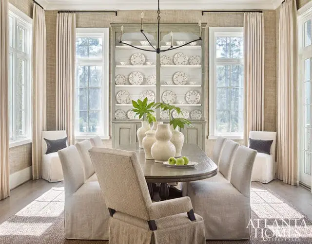 Neutral dining room with oval wood table, slipcovered chairs, textured wallpaper, and a tall gray hutch filled with decorative plates.