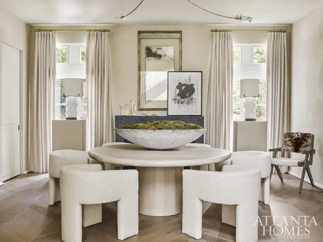Dining room with a round light wood table surrounded by four white bouclé stools and a chair with cowhide upholstery. A large stone bowl filled with moss sits on the table.