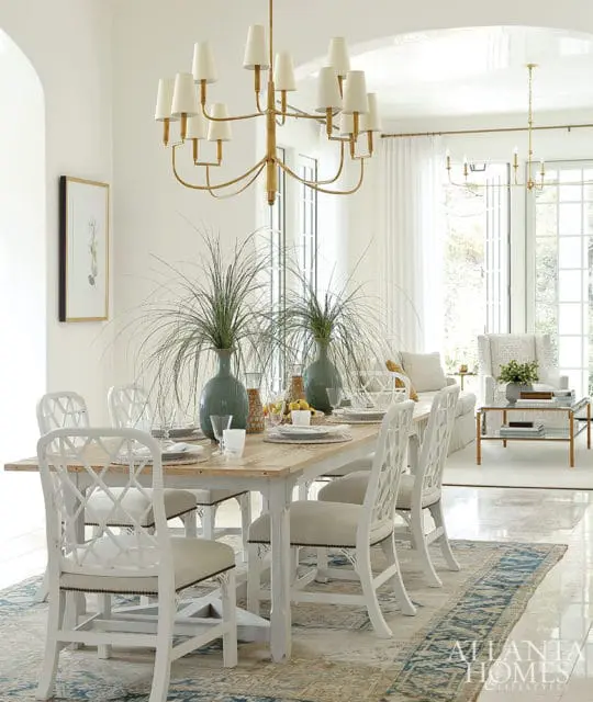 Dining room with a light wood table and white lattice-back chairs, set with green vases of grass and a patterned rug.