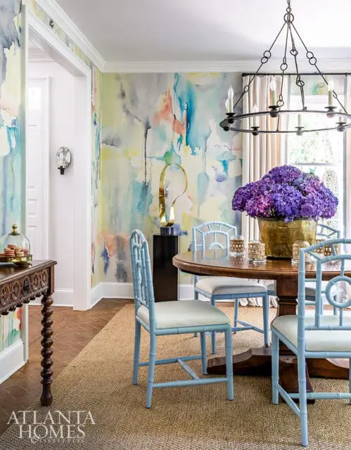 Dining room with abstract watercolor wallpaper, light blue bamboo-style chairs with cream cushions, and a round wooden table topped with a large vase of purple hydrangeas.