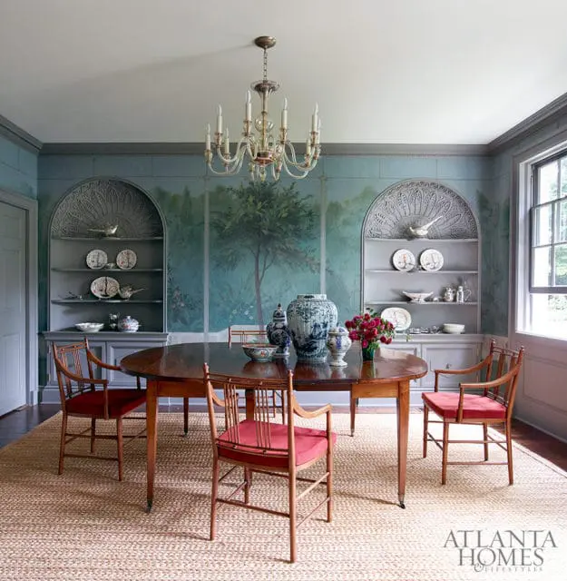 Dining room with oval wood table, red upholstered chairs, and walls featuring a mural of a tree and landscape. Built-in shelves display blue and white porcelain.