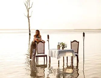 Woman in a shimmering dress stands by a table set for two in shallow water, with tiki torches on either side and a bare tree in the background.
