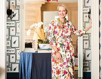 Woman in a floral dress stands in a doorway with patterned wallpaper featuring framed pictures of birds and butterflies.