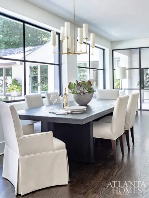 Dining room with a dark wood table, upholstered chairs, and a modern gold chandelier. Large windows overlook an outdoor area.