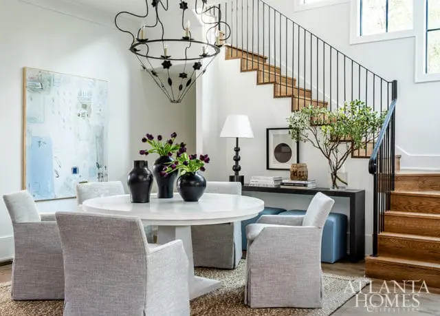 Dining area with a white round table, upholstered chairs, black vases with purple tulips, abstract art, and a black metal chandelier. A wooden staircase with black railing is visible in the background.
