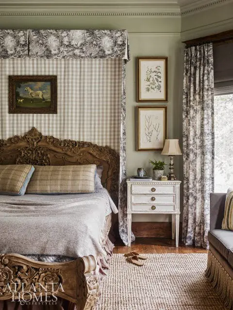 Bedroom with a carved wooden bed, plaid and toile patterned canopy and curtains, and two framed botanical prints on a sage green wall.