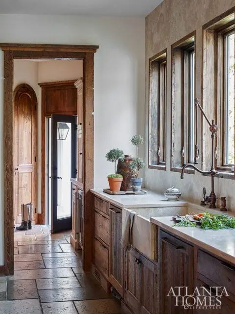 Rustic kitchen with reclaimed wood cabinetry, concrete countertops, apron sink, and a view into a hallway with arched doorways and stone flooring.