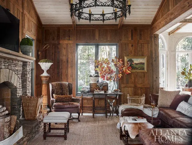 Rustic living room with wood-paneled walls, stone fireplace, leather sectional sofa, and antler chandelier. A deer statue stands near a table with autumn foliage.